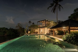 A nighttime view of a modern resort with a swimming pool, a covered outdoor area, and lush tropical vegetation.