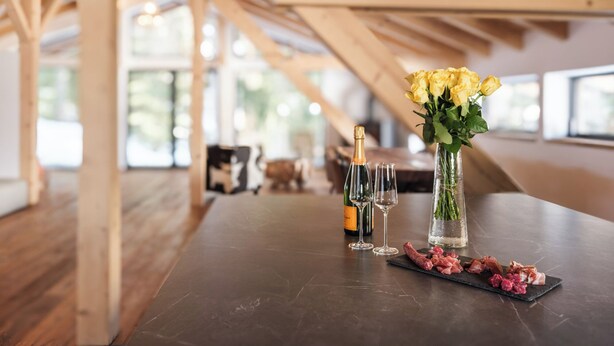 A dining table with a bottle of champagne, two wine glasses, a vase with yellow flowers, and a selection of meats on a slate board.