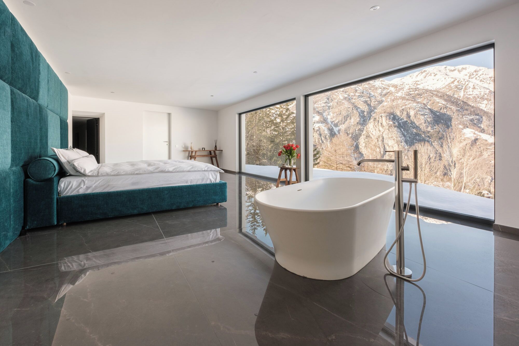 Ein modernes Schlafzimmer mit einem großen Bett, einer freistehenden Badewanne und Bergblick durch große Fenster.