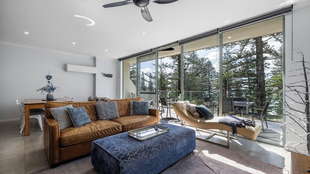 Un salon moderne avec un canapé en cuir marron, un pouf bleu et une table basse en verre. On y trouve également un ventilateur de plafond, un coin repas avec une table et des chaises en bois, ainsi qu’une grande porte-fenêtre coulissante donnant sur un balcon avec vue sur les arbres.