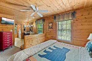 Quarto de cabana de madeira com cama, cômoda vermelha, ventilador de teto e banheiro com pia e espelho.