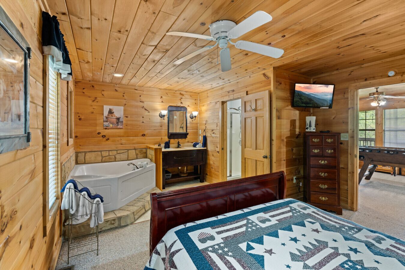 A wooden cabin bedroom with a large bed, a wooden dresser, a vanity with a mirror, a bathtub, and a ceiling fan.