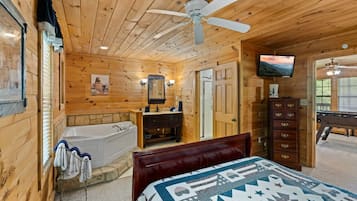 A wooden cabin bedroom with a large bed, a wooden dresser, a vanity with a mirror, a bathtub, and a ceiling fan.