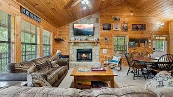 A cozy cabin interior with a stone fireplace, wooden walls, a dining area, and a couch.