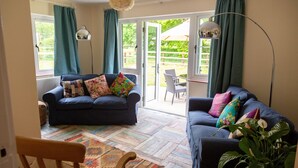 A cozy living room with a blue sofa, colorful cushions, a patterned rug, and a view of the garden through open doors. - 3 Bedroom idyllic countryside cottage in the middle of rural Wiltshire (Coate)