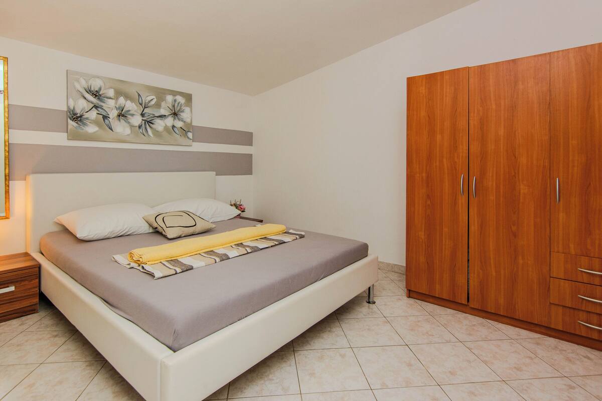 A bedroom with a bed, a wooden wardrobe, a painting on the wall, and a nightstand.
