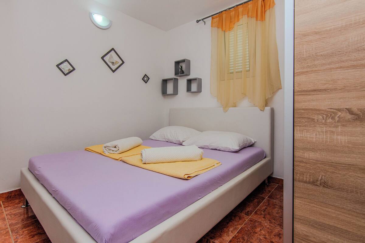 A neatly made bed with a purple bedspread, white pillows, and two towels. There are wall-mounted shelves with decorative items and a window with orange curtains.