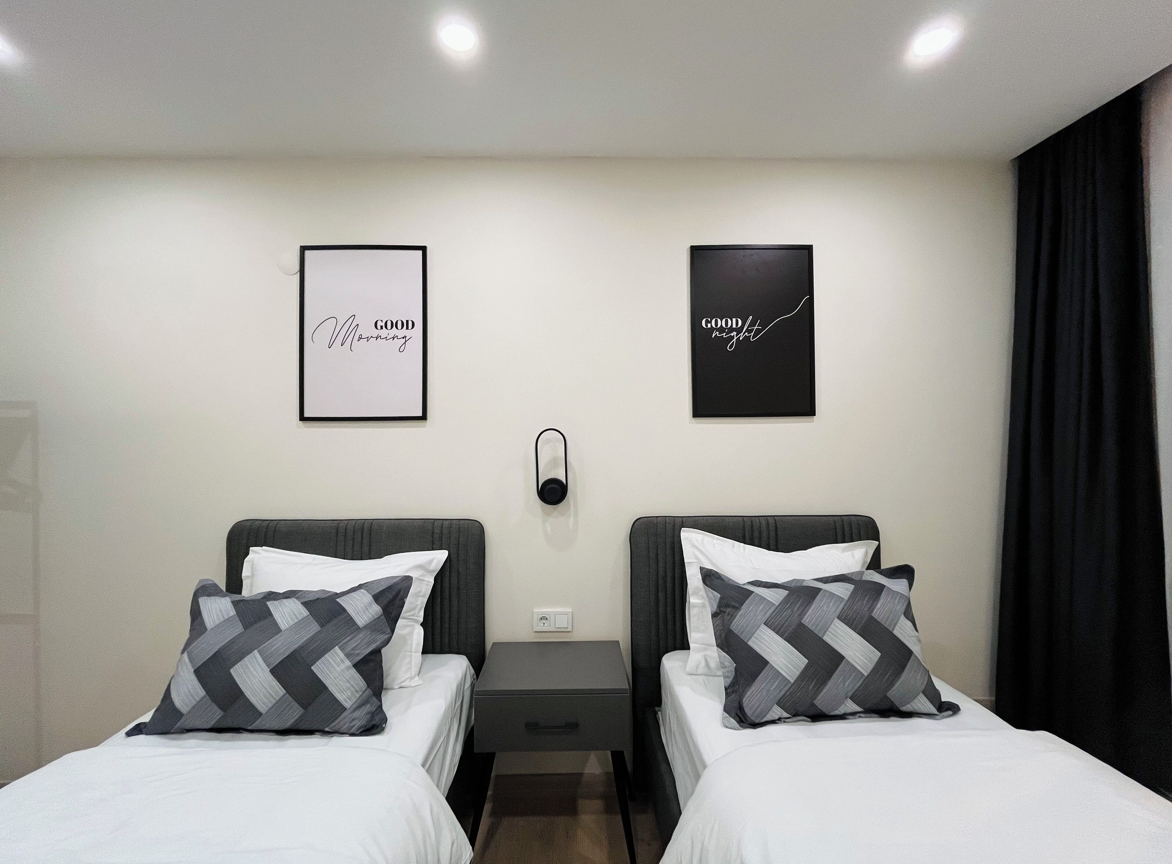 Ett hotellrum med två sängar, var och en med en grå och vit kudde med sicksackmönster, ett litet sängbord och en väggmonterad lampa. På väggen hänger två inramade tavlor, den ena med texten ”God morgon” och den andra med texten ”God natt”.
