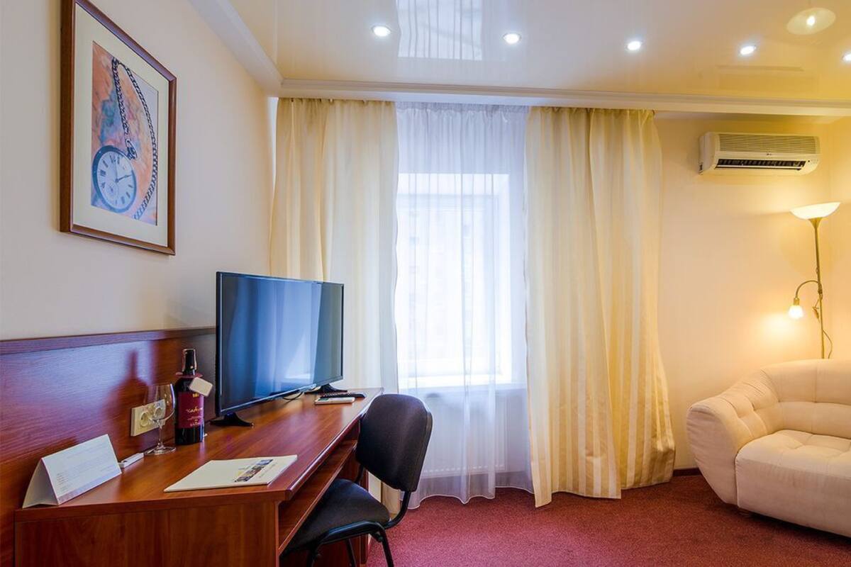 Ein Hotelzimmer mit Schreibtisch aus Holz, Fernseher, einer Couch und einem großen Fenster mit Vorhängen.
