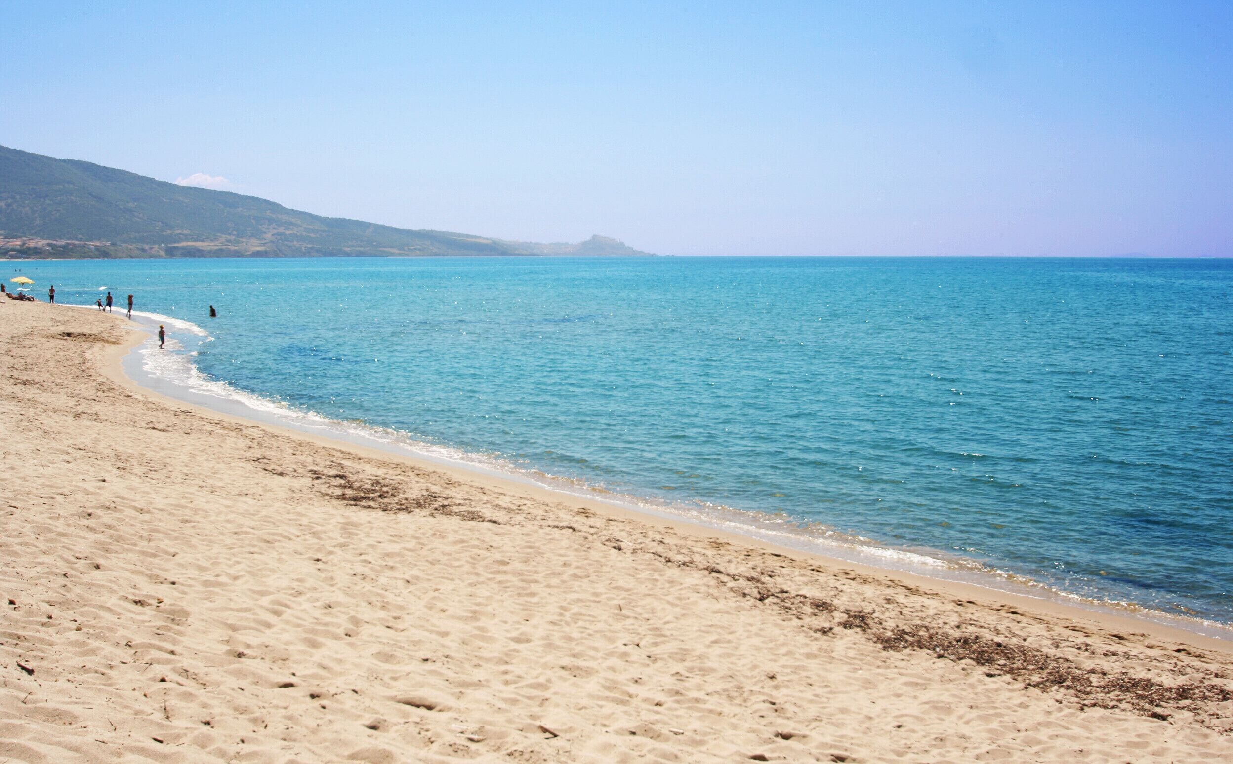 Uma praia com águas azuis cristalinas, areal arenoso e algumas pessoas caminhando.