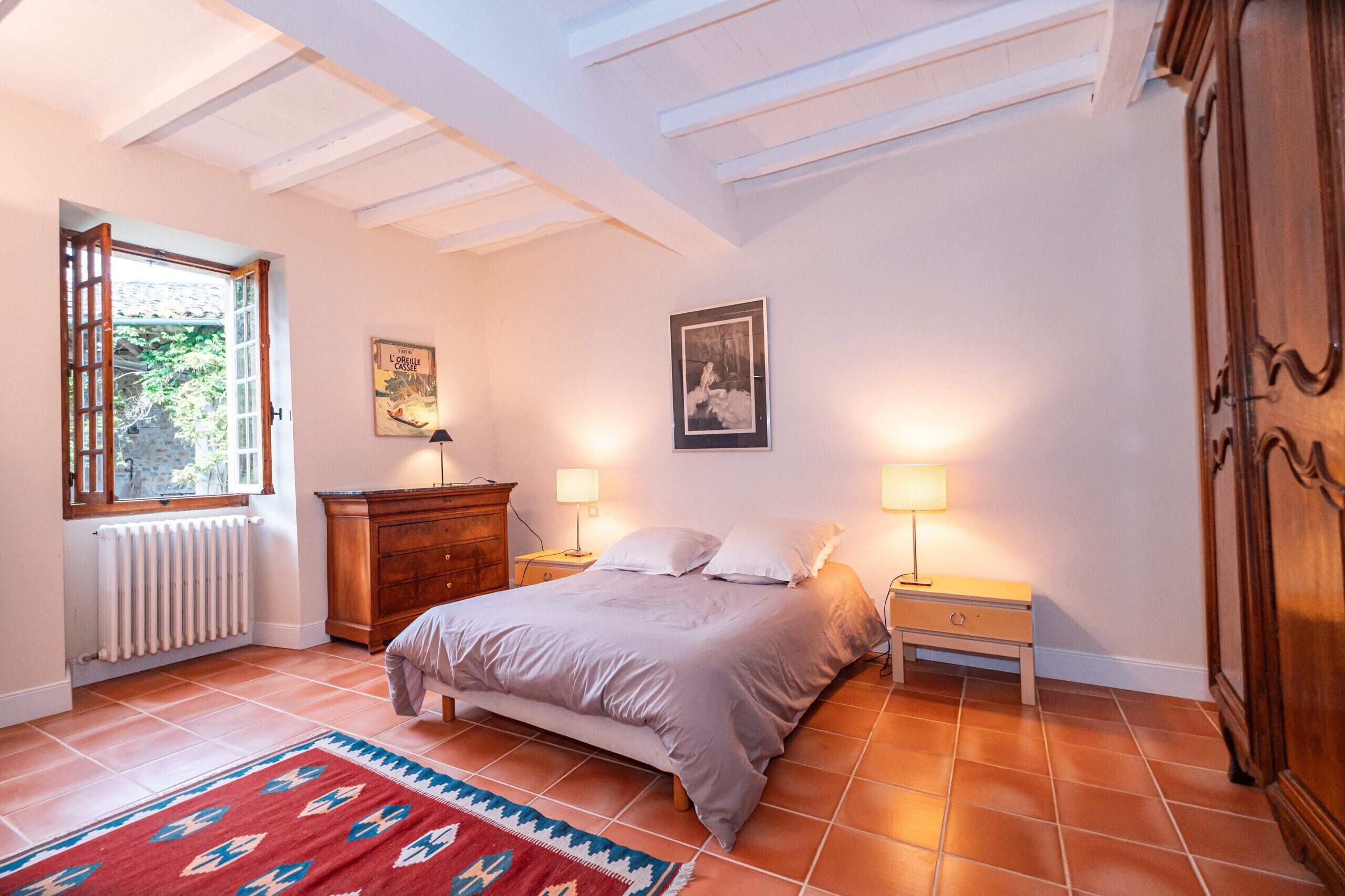A bedroom with a bed, two bedside tables with lamps, a wooden wardrobe, a radiator, and a window with a view of greenery.