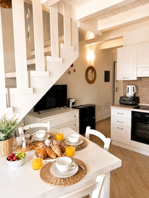 Una cocina moderna con muebles blancos, un comedor con una mesa para el desayuno que incluye cruasanes y jugo de naranja, y una escalera.