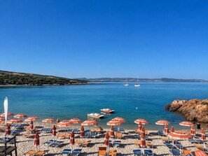 Uma praia com fileiras de espreguiçadeiras e guarda-sóis, um litoral rochoso e veleiros na água.