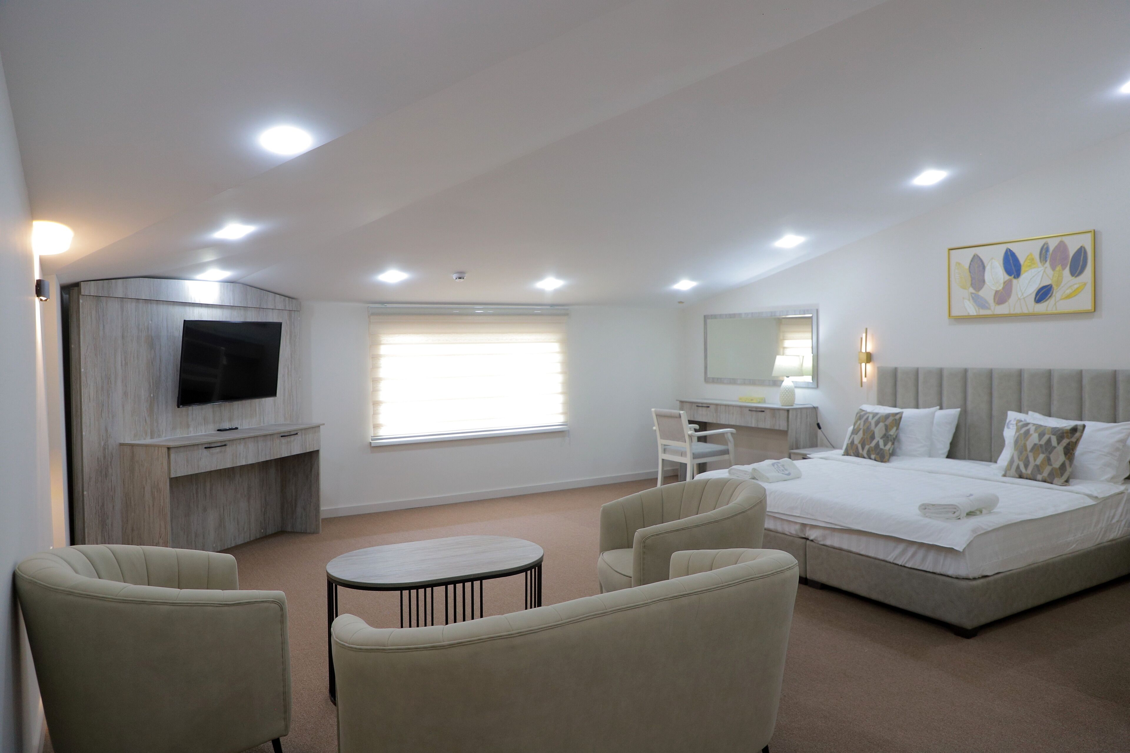 A modern hotel room with a large bed, a desk, a flat-screen TV mounted on a wooden panel, a round coffee table, and two armchairs.