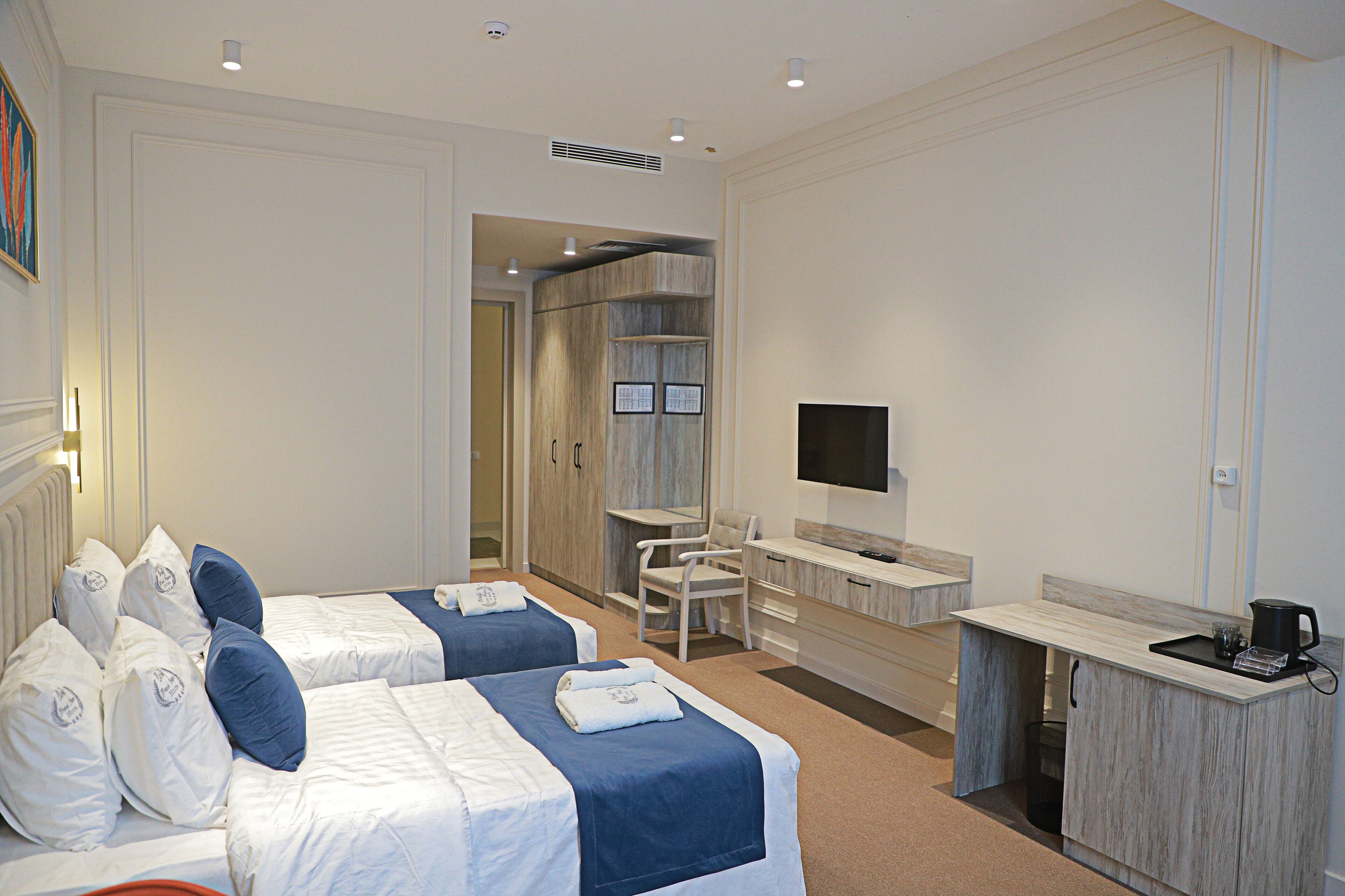 A hotel room with two beds, a desk, a chair, a TV, and a small kitchenette.