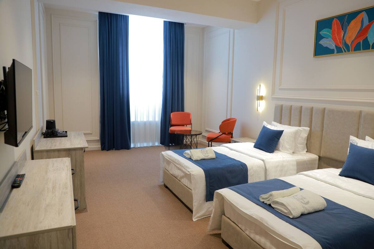 A hotel room with two beds, a desk, a chair, a TV, and a large window with curtains.