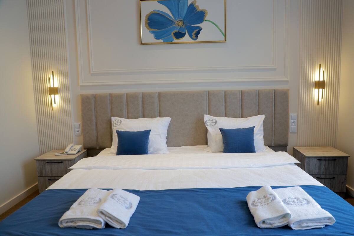 A neatly made bed with blue and white bedding, two bedside tables with lamps, and a framed floral artwork on the wall.