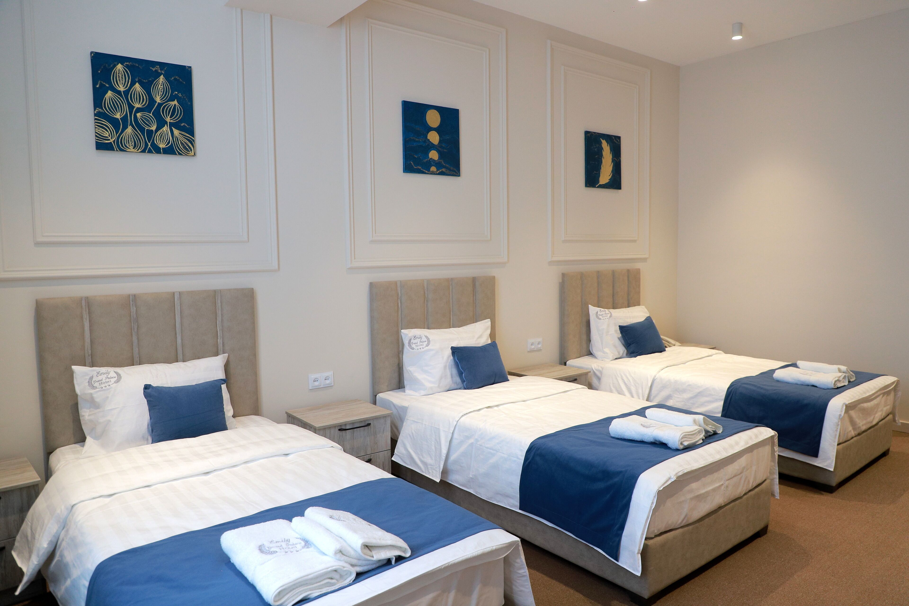 A hotel room with three beds, each with a blue and white bedspread, and artwork on the walls.