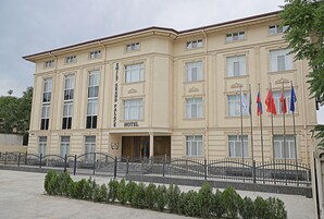 A large, cream-colored building with a sign reading "EMILY GRAND PALACE HOTEL." It features multiple windows, a black iron fence, and several flags in front.