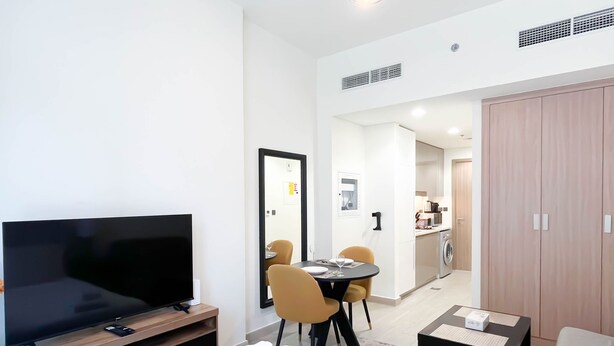 A modern hotel room with a flat-screen TV, a dining area with a table and chairs, a wardrobe, and a small seating area with a sofa and a lamp. - Urban Charm: Studio With Balcony in Azizi Riviera (Dubai)