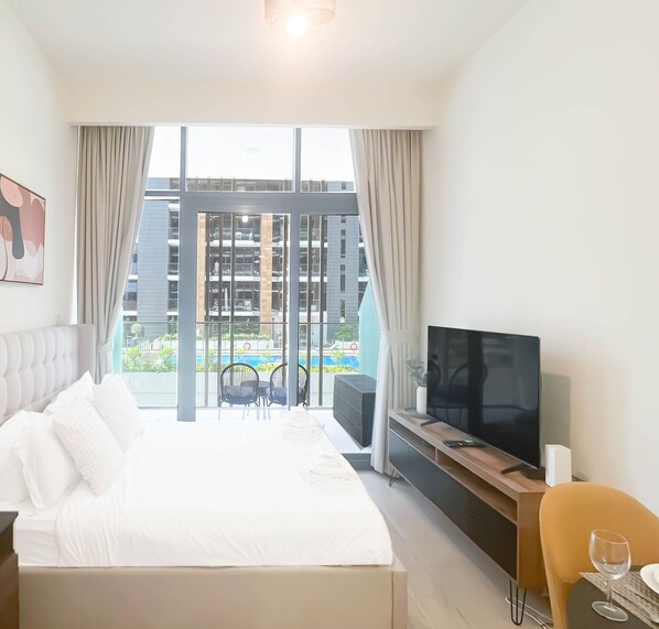 A modern hotel room with a large bed, a TV, a desk, and a view of the cityscape. - Urban Charm: Studio With Balcony in Azizi Riviera (Dubai)