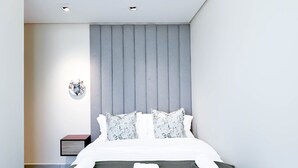 A modern bedroom with a large bed, grey headboard, white bedding, and a small bedside table with a lamp. - Stylish 1BR With Balcony City Views O2 Tower, JVC (Dubai)
