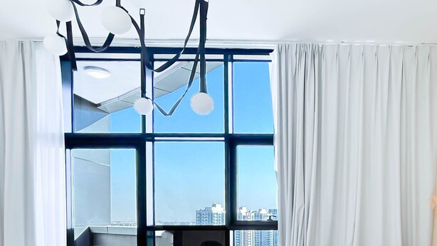 A modern living room with a large window, a sofa, a round coffee table, and a hanging light fixture. - Stylish 1BR With Balcony City Views O2 Tower, JVC (Dubai)