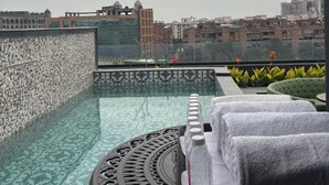 Uma piscina infinita no terraço com piso estampado, uma mesa redonda com estrutura metálica decorativa e vista para a paisagem urbana.