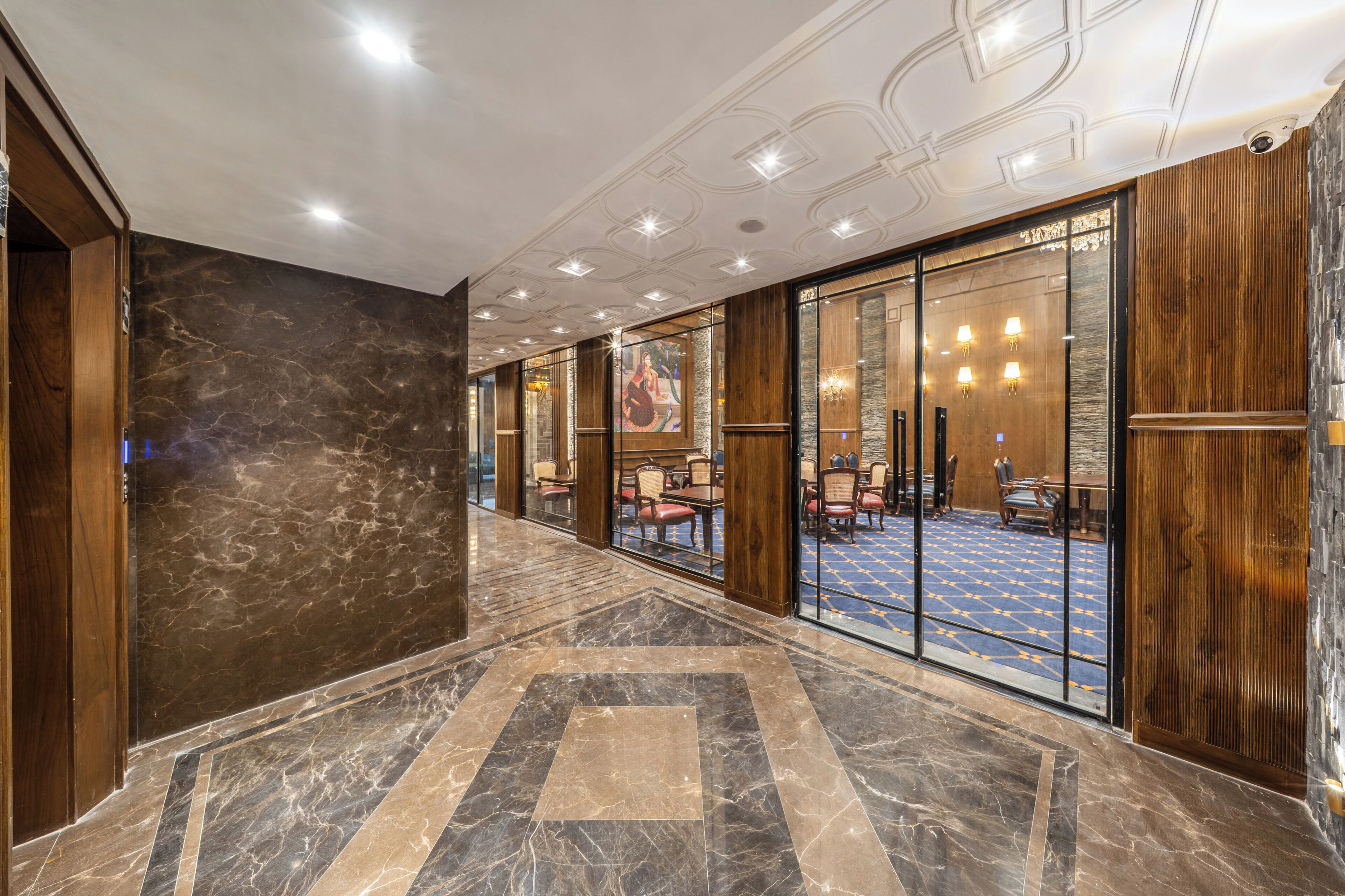 A hotel lobby with marble flooring, wooden paneling, and a blue carpeted area with tables and chairs.