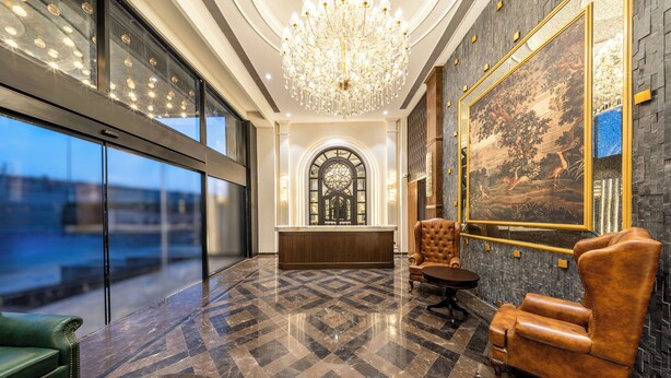 Eine Lobby mit einer Rezeption, einer großen Kristallleuchtre, einem grünen Sofa, einem braunen Ledersessel und einem großen Gemälde an der Wand.