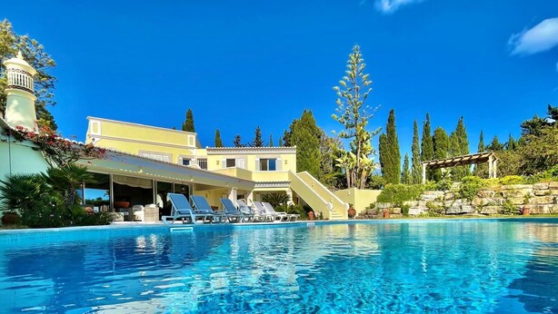 A swimming pool with lounge chairs, a covered area, and a building with a light-colored facade and a tower.