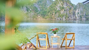 A wooden deck with two chairs and a small table, overlooking a calm lake with a mountainous backdrop. - Lan Ha Floating Homestay (Hai Phong)