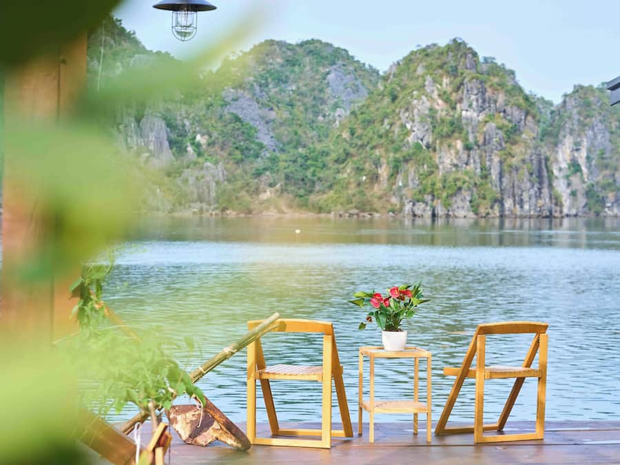 Eine Holzterrasse mit zwei Stühlen und einem kleinen Tisch, mit Blick auf einen stillen See vor einer Bergkulisse.