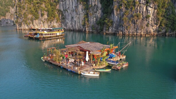 A floating structure with a covered deck, a small boat docked nearby, and a large boat in the background. - Lan Ha Floating Homestay (Hai Phong)