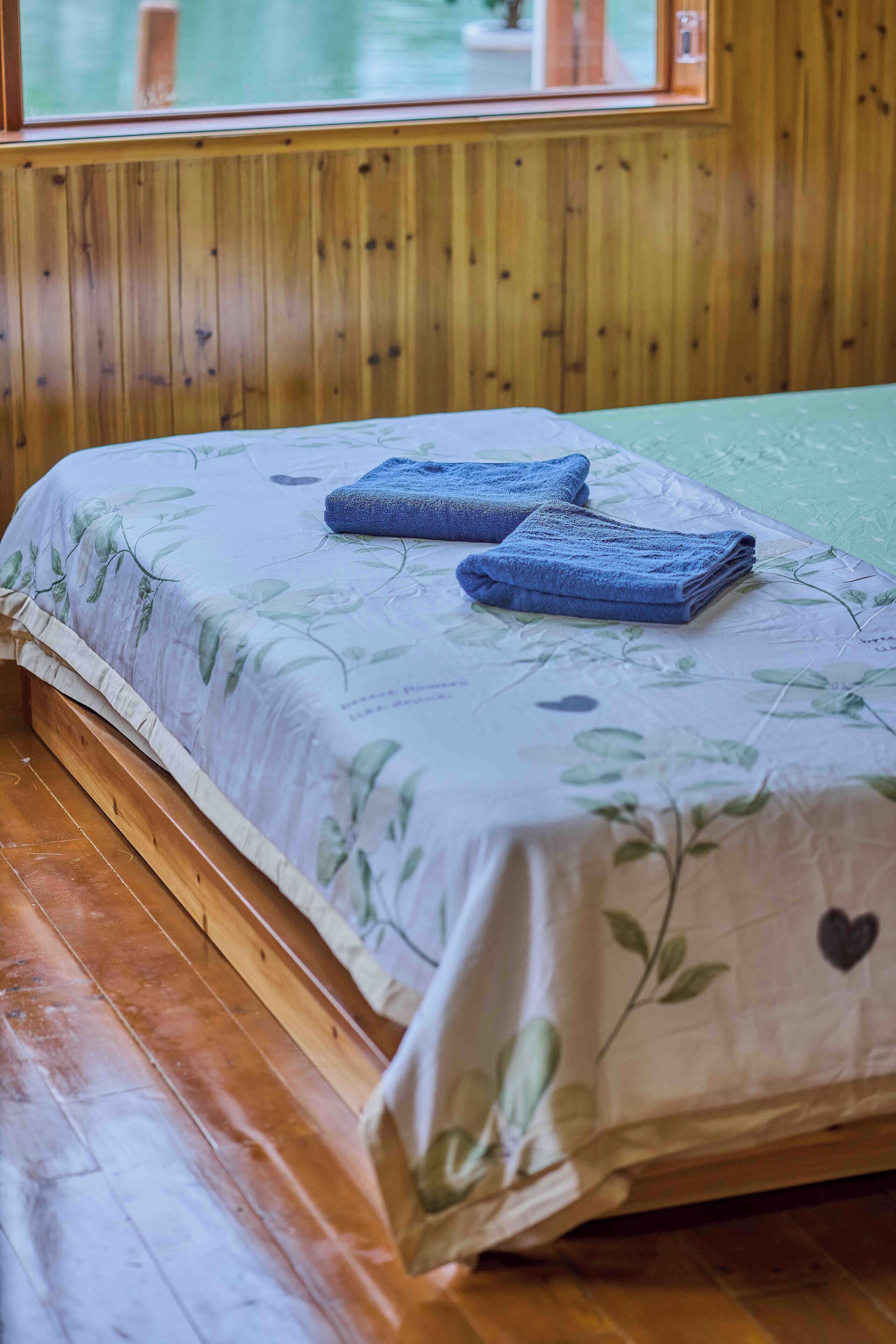 A wooden paneled room with a bed featuring a floral bedspread and blue towels.