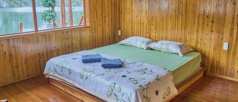 A wooden-paneled room with a bed, a window overlooking a body of water, and a view of trees and a ladder outside.