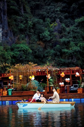 A boat with people on it, a wooden structure with plants in the background, and a forested area. - Lan Ha Floating Homestay (Hai Phong)
