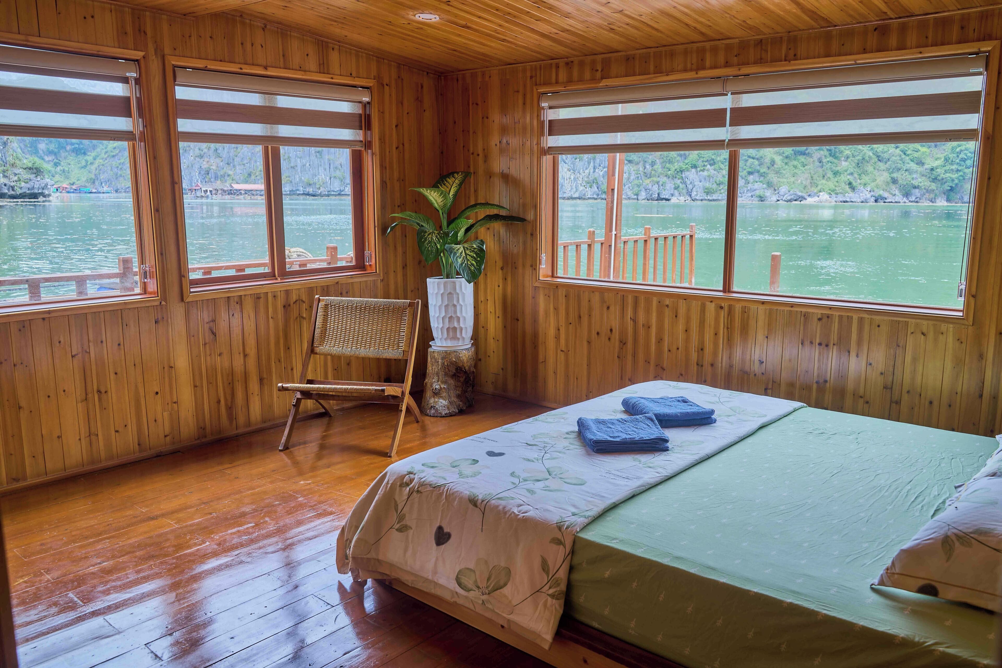 A wooden cabin room with a bed, a chair, a potted plant, and large windows overlooking a body of water.