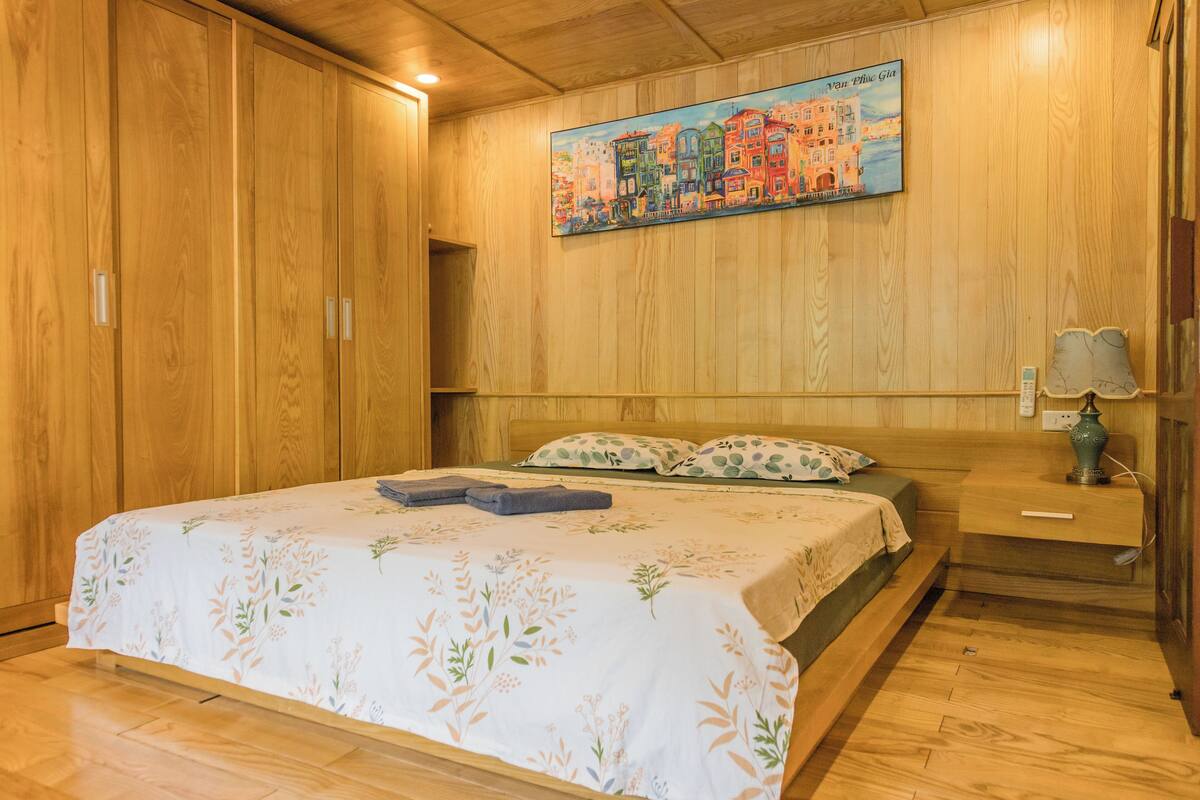 A wooden-paneled room with a bed, a nightstand, a lamp, and a painting of a European town.