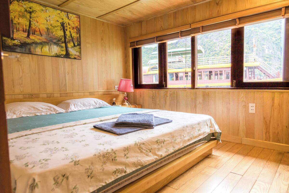 A wooden room with a bed, a painting, a window with a view of a train, and a lamp.