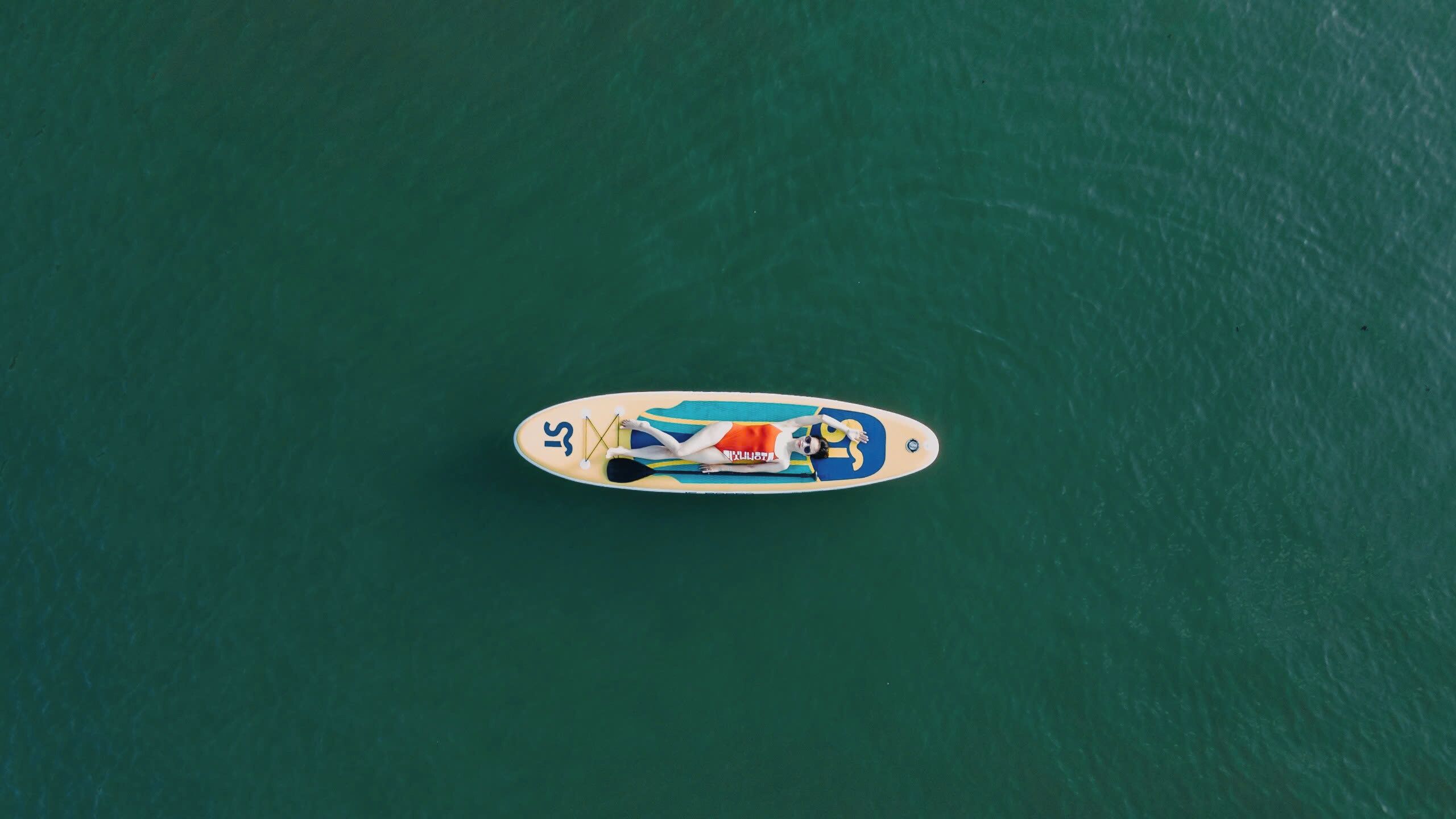 Luftaufnahme eines Stand-Up-Paddleboards, auf dem sich eine Person entspannt treibt, inmitten von grünem Wasser.