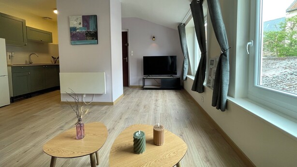 Un salon moderne avec un téléviseur à écran plat, deux petites tables rondes en bois, un tableau accroché au mur et une vue sur l’extérieur grâce à une grande fenêtre.