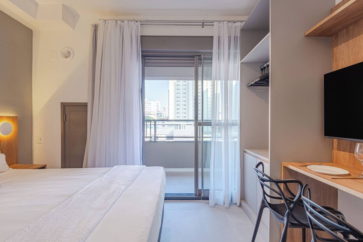 Ein Hotelzimmer mit Bett, Schreibtisch mit Stuhl, Fernseher und einem Balkon mit Vorhängen.