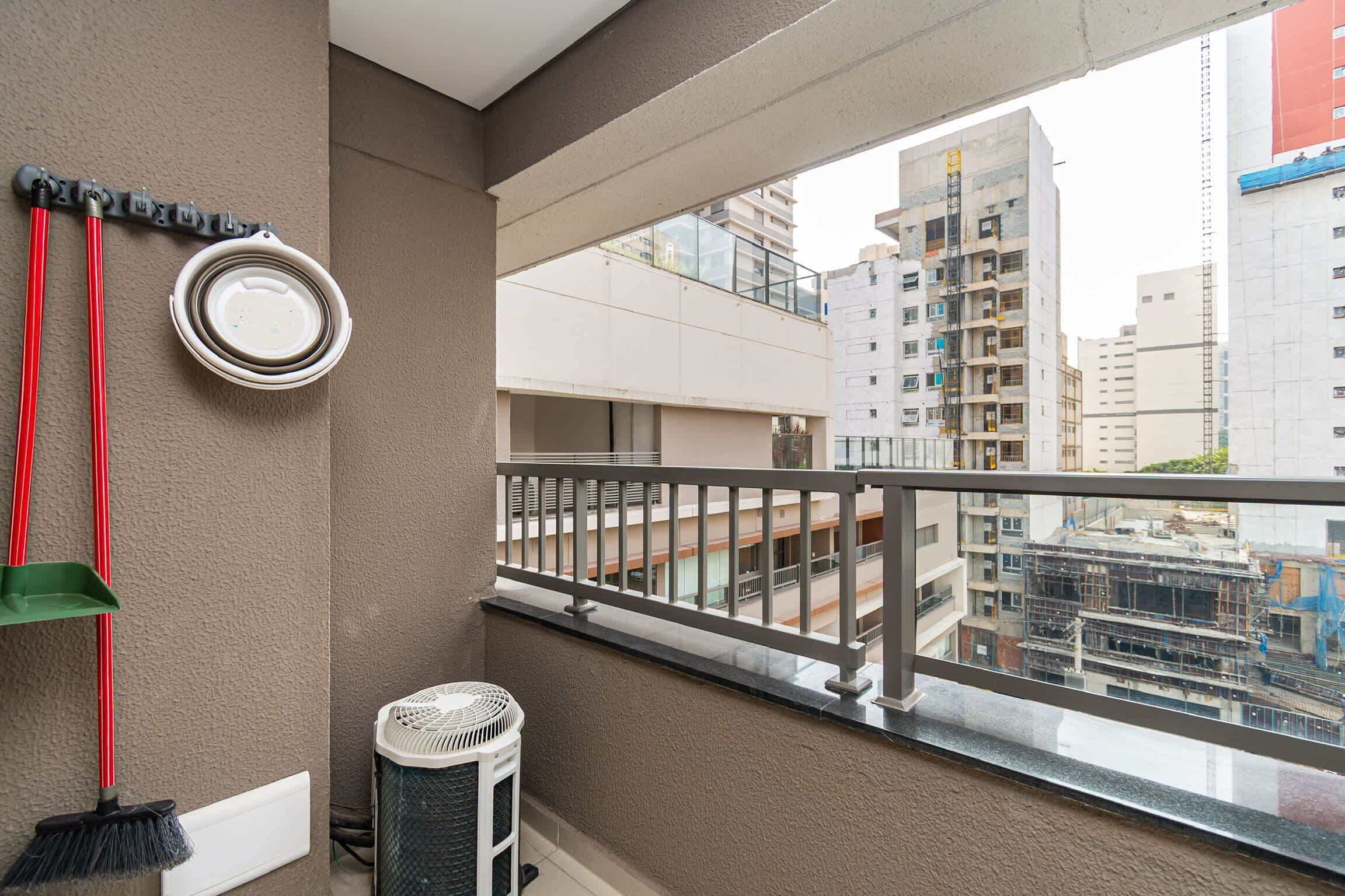 A balcony with a view of surrounding buildings, a wall-mounted air conditioning unit, and a broom leaning against the wall.