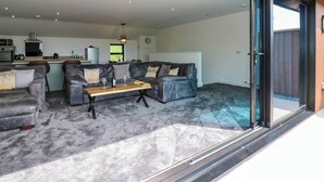 A modern living room with a grey sofa set, a wooden coffee table, and a glass sliding door leading to an outdoor area. - Sycamore Cottage (Frizington)