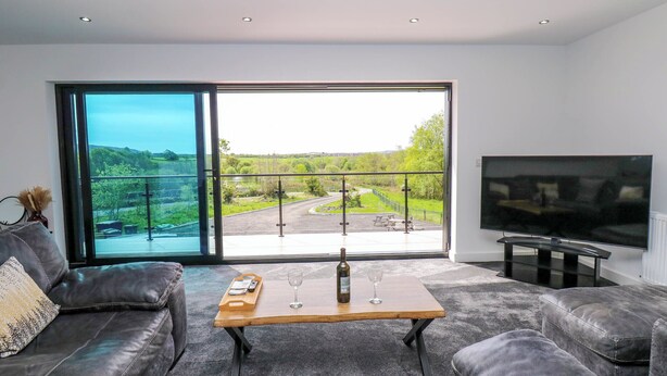 A modern living room with a leather sofa, a wooden coffee table with a bottle and glasses, and a large TV. - Sycamore Cottage (Frizington)