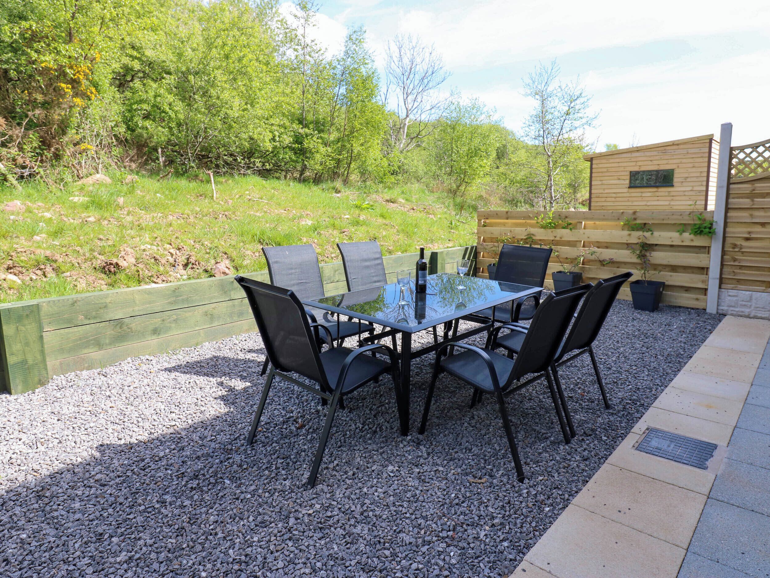 A glass-top table with six chairs, surrounded by gravel, with a wooden fence and greenery in the background.
