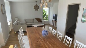 A dining area with a wooden table surrounded by white chairs, a sofa, and a television in the background. - New, seaside house, Beg meil, for groups and families  (Fouesnant)