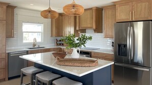 A modern kitchen with wooden cabinets, a stainless steel refrigerator, a large island with stools, and a hanging light fixture. - 3+ BR Beach ViewHouse (Palm Coast)