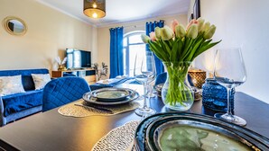 Un coin repas avec une table dressée pour deux personnes, un canapé bleu, un téléviseur et une fenêtre avec des rideaux.